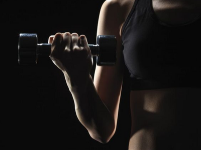 A closeup of a woman holding a small dumbbell against a black background in low lighting